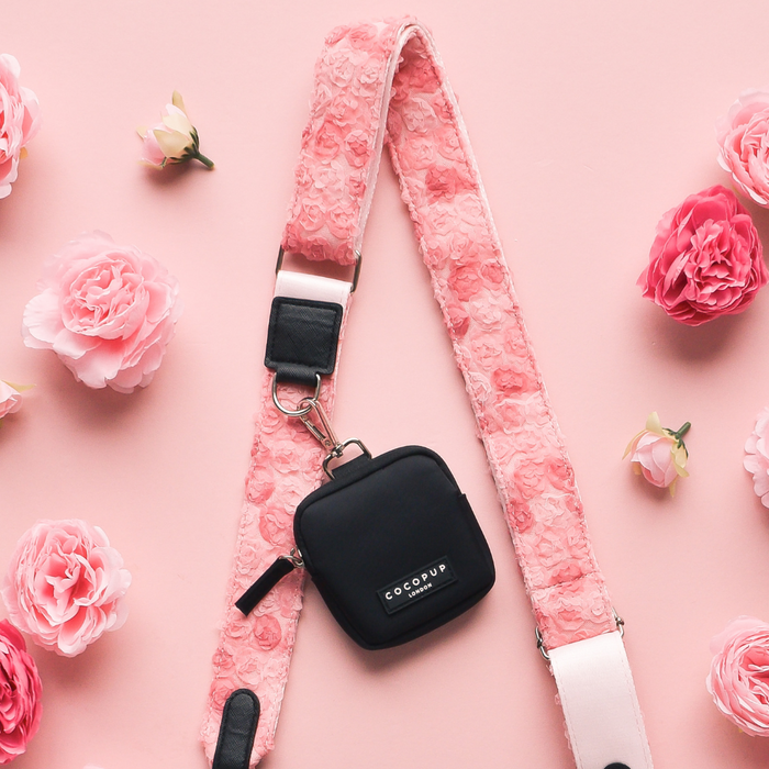 Cocopup London Peony Pup bag strap with black case on pink background, surrounded by pink artificial flowers.
