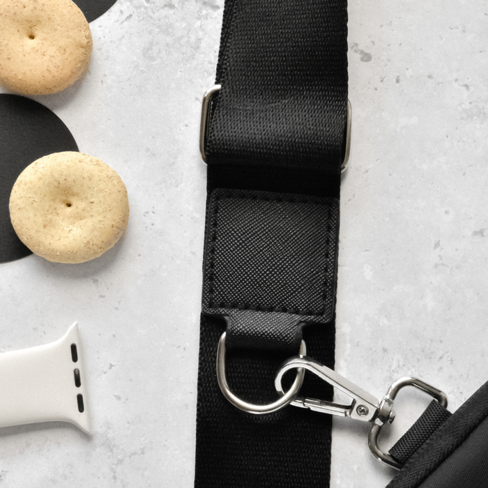 A close-up of the Cocopup London Bag Strap - Plain Black, featuring an adjustable design and a silver hook clasp, lying on a light gray surface. To the left, there are gingernut cookies and a white watch strap. The scene is minimalistic with items neatly arranged.