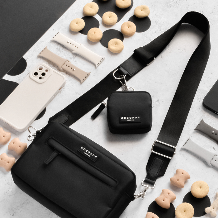 A flat lay photo showcases a black crossbody bag with an adjustable, plain black dog walking bag strap (Product Name: Bag Strap - Plain Black) from Cocopup London. Alongside it is a small black pouch featuring "COCOPUP" branding. The scene is set with dog treat biscuits, interchangeable watch straps in neutral colors, and a white smartphone encased in beige on a light gray surface.