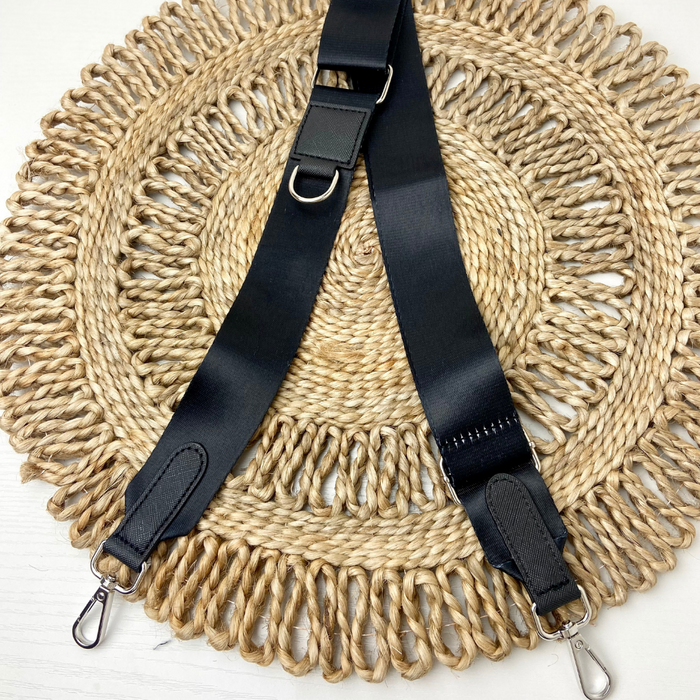 A Cocopup London Bag Strap in plain black polyester, featuring adjustable length and metal clips at both ends, is laid out on a circular woven straw placemat with intricate patterns. The strap includes a metal D-ring attached to a leather patch for added functionality.
