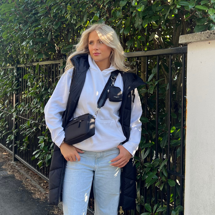 A person with long blonde hair stands in front of a fence and greenery, wearing a white hoodie, light blue jeans, and a black puffy vest. They carry an adjustable Bag Strap - Plain Black by Cocopup London, paired with a small black crossbody bag and matching pouch. They're looking off to the side with a relaxed posture.
