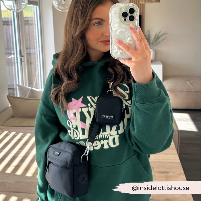 A woman stands in a well-lit room taking a mirror selfie. She wears a dark green hoodie with colorful text and has two black crossbody bags, one featuring an adjustable Cocopup London Bag Strap - Plain Black. Her phone case is clear with a white pattern. The Instagram handle @insidelottishouse is visible.
