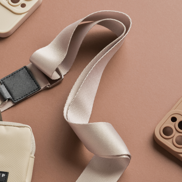 Close-up of a Bag Strap - Plain Nude from Cocopup London and light brown phone cases on a brown-colored surface. The camera strap, an everyday essential made of fabric, includes a black adjustment clip. The scene is well-lit, focusing on the timeless colours and textures of the objects.