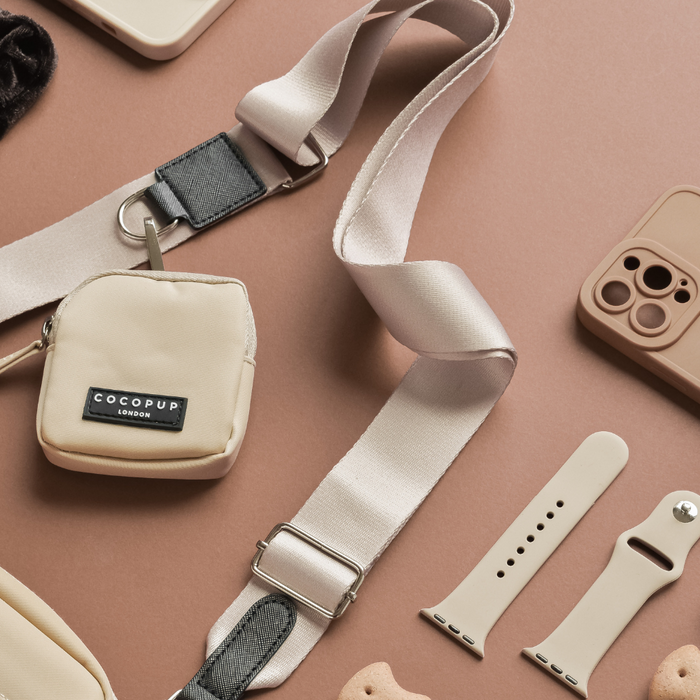 A plain nude crossbody strap, a mini pouch labeled "COCOPUP LONDON," beige Apple Watch bands, a taupe iPhone case with a paw print cutout, and a beige bone-shaped dog treat are arranged on a peach-colored background, showcasing timeless everyday essentials perfect for your dog walking bag.