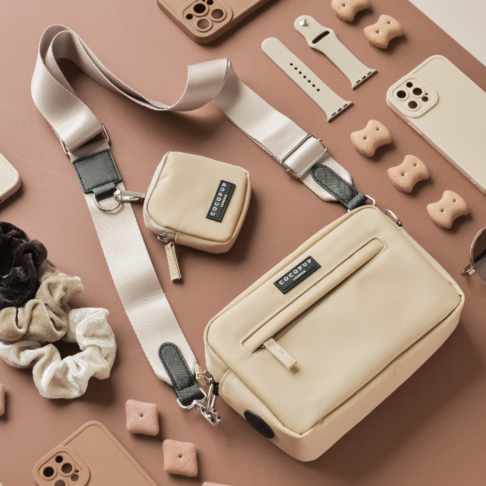 A neatly arranged flat lay of timeless beige and tan accessories, featuring a Cocopup London crossbody dog walking bag with a matching smaller pouch, phone cases, a watch band, hair scrunchies, and dog treats on a pinkish-tan surface. The bag has the "Bag Strap - Plain Nude" strap with black accents and is labeled "COCOWAY".