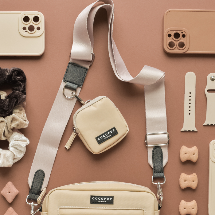 An assortment of beige and cream everyday essentials neatly arranged on a brown background. Items include a crossbody dog walking bag, a small pouch, phone case, smartwatch strap, scrunchies, and dog treats—all in timeless colors. The bag and pouch both feature "Cocopup London" branding along with the Bag Strap - Plain Nude.