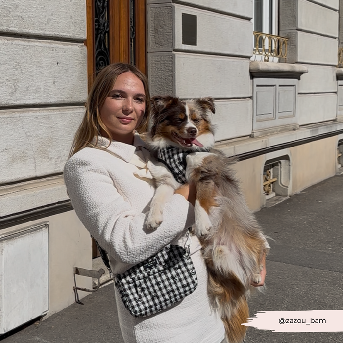 Woman in white with a Cocopup London pink patterned bag strap, holding a small dog on a sidewalk by a stone building.
