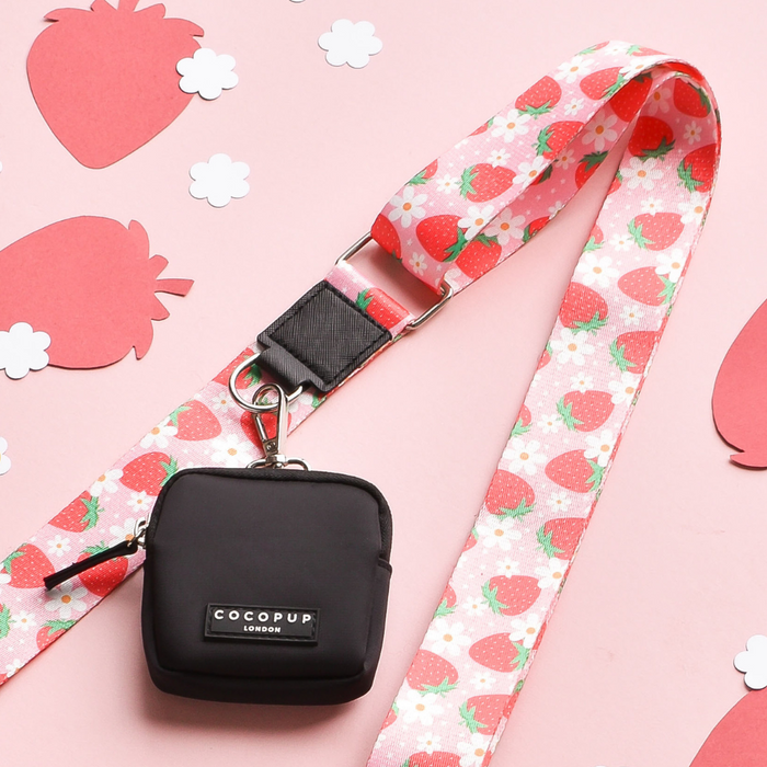 A chic square pouch in black, featuring a silver clip and a "Cocopup London" label, dangles from the vibrant Pupberry Fields bag strap. This lanyard is beautifully decorated with colorful strawberries and flowers. Placed on a light pink surface adorned with paper strawberry cutouts, this accessory is perfect for stylish dog walks.