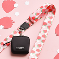A chic square pouch in black, featuring a silver clip and a "Cocopup London" label, dangles from the vibrant Pupberry Fields bag strap. This lanyard is beautifully decorated with colorful strawberries and flowers. Placed on a light pink surface adorned with paper strawberry cutouts, this accessory is perfect for stylish dog walks.