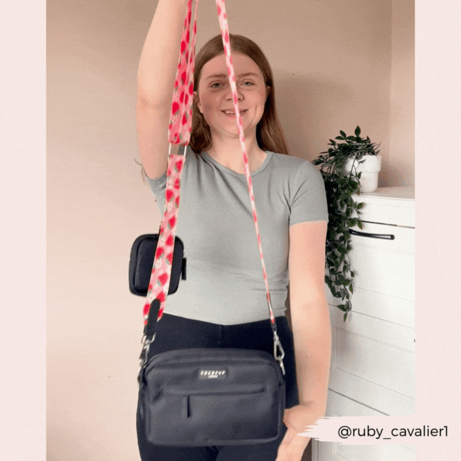 A woman with long hair is smiling while holding a black crossbody bag featuring the Bag Strap - Pupberry Fields by Cocopup London. She stands in front of a light wall adorned with a white chest of drawers and a plant. The Instagram handle @ruby_cavalier1 is visible in the corner, hinting at her chic, stylish dog walks.