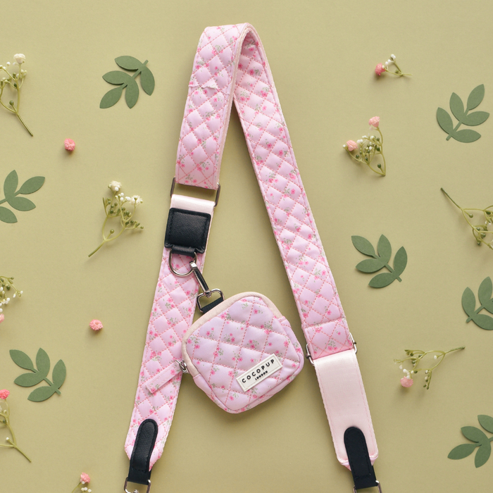 Pink quilted floral bag strap with a small matching pouch, displayed among green leaves and tiny white flowers.