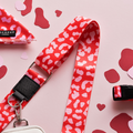 A pink lanyard with a red and white camouflage pattern is laid on a pink surface. Around it, there are scattered red and pink heart-shaped confetti pieces. Part of a pink cloth with a label reading "PUP LONDON" is also visible, next to what seems like the Bag Strap - Red & Pink Leopard by Cocopup London — ideal for your dog walking accessory needs.