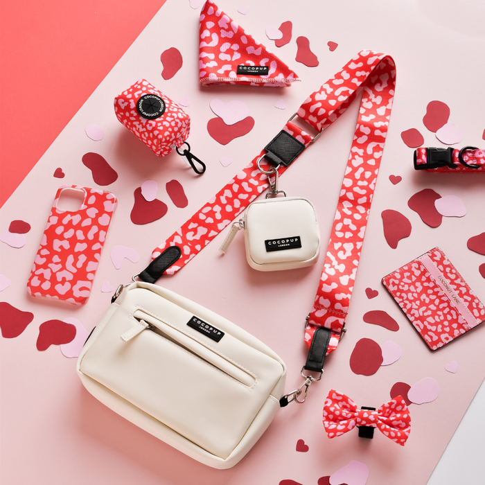 An arrangement of white and red accessories against a pink and red background. There is a white crossbody bag featuring the Cocopup London Bag Strap - Red & Pink Leopard, a red keychain pouch, a patterned phone case, a bow, a wallet, and other small items. Hearts and confetti are scattered around.
