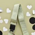 A pale green adjustable strap adorned with heart patterns is laid flat on a light green background. Surrounding the Luxe Bag Strap - Sage Heart by Cocopup London are scattered paper cutouts in the shapes of hearts and circles, with hearts in white and circles in black.