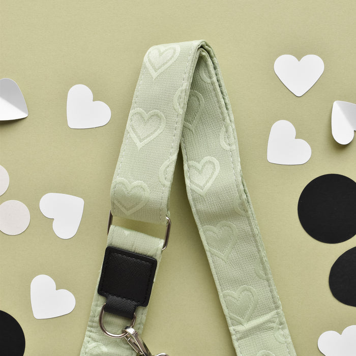 A pale green adjustable strap adorned with heart patterns is laid flat on a light green background. Surrounding the Luxe Bag Strap - Sage Heart by Cocopup London are scattered paper cutouts in the shapes of hearts and circles, with hearts in white and circles in black.
