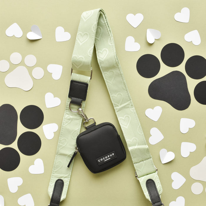 The Luxe Bag Strap - Sage Heart from Cocopup London, featuring a green design with white heart patterns and a small black pouch attached, rests on a light green surface adorned with black dog paw prints and white heart-shaped cutouts. The adjustable strap ensures comfort, making it perfect for your Cocopup Dog Walking Bag.