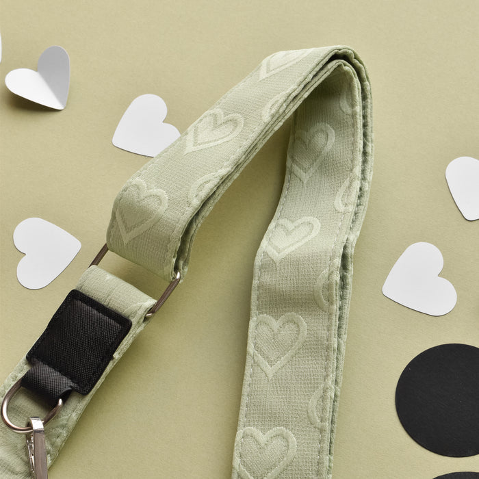 A light green lanyard with a pattern of embossed hearts lies on a beige surface. Surrounding the Luxe Bag Strap - Sage Heart from Cocopup London are scattered white paper hearts and two black circular cutouts. A metal clip is visible at the end of the adjustable strap.