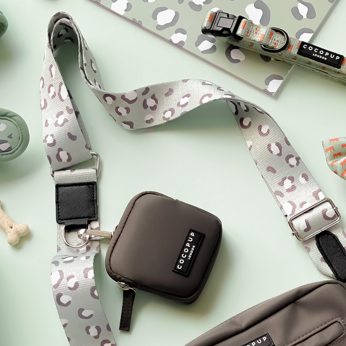 Image of various dog accessories on a light green surface. Items include a leopard print dog leash titled "Bag Strap - Sage Leopard" from Cocopup London, a small gray pouch with the "Cocopup London" label, a matching waste bag holder, a dog collar, a green retractable leash, and a bone-shaped dog treat.