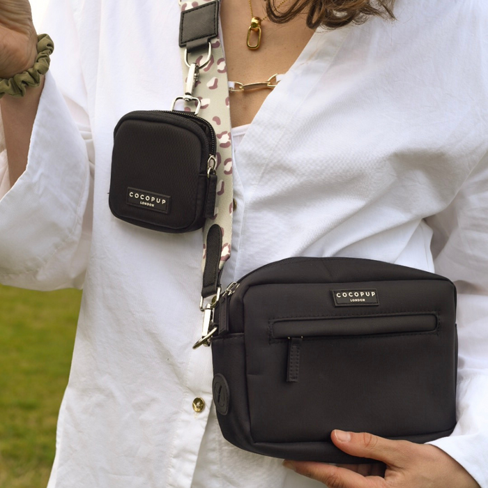 A person wearing a white shirt is seen outdoors, holding a black Cocopup London Dog Walking Bag from the Sage Leopard Collection with a "COCUP LONDON" label. They also have a smaller black pouch attached to the Sage Leopard Bag Strap. A green hair tie is on their wrist.