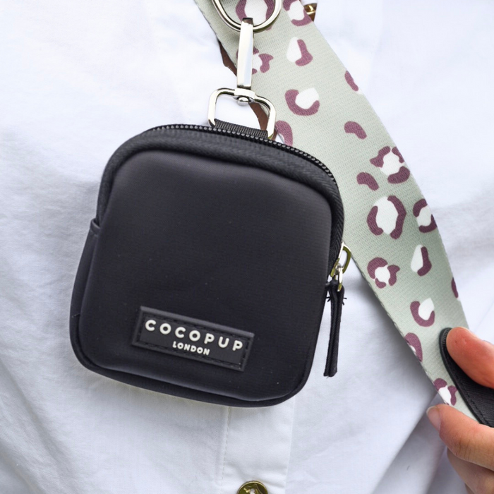 A close-up of a small black zippered pouch with "Cocopup London" in white letters on the front. The pouch is attached to the Bag Strap - Sage Leopard from the Cocopup Dog Walking Bag collection, worn over a white shirt. A hand is visible adjusting the strap.