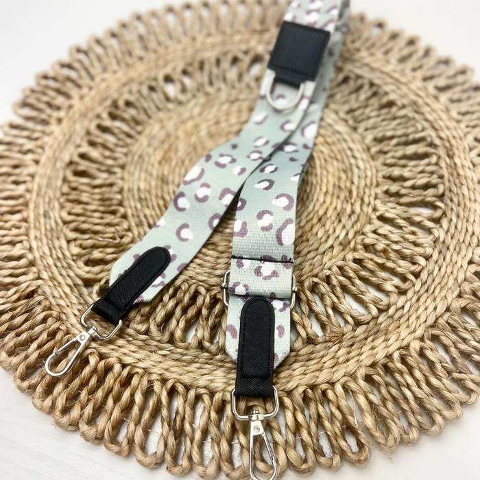 The Bag Strap - Sage Leopard from Cocopup London, showcasing a light-colored base with a leopard print pattern, is placed on a round, woven placemat. This detachable strap is part of the Cocopup Dog Walking Bag collection and features silver clasps and black leather-like accents at its ends.