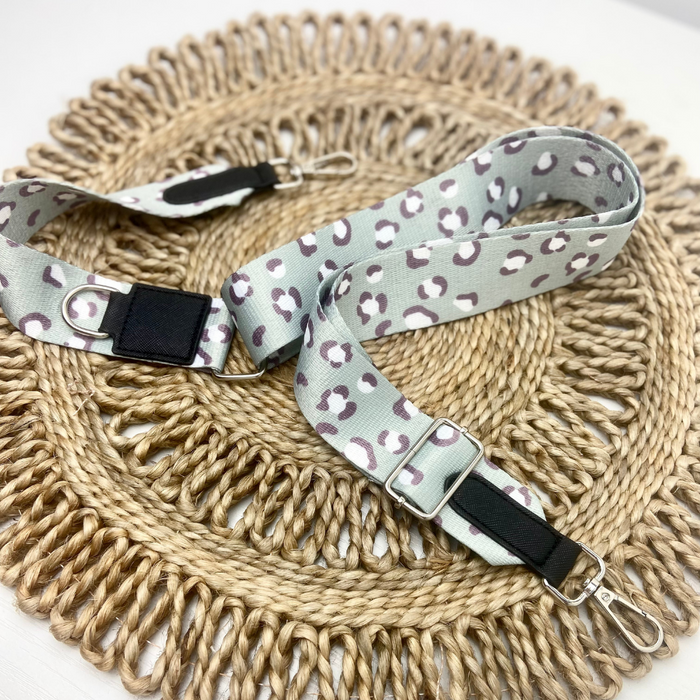 A Bag Strap - Sage Leopard from Cocopup London lies on a circular woven mat. This light gray strap with a white and brown leopard print design features chic black adjustable buckles and metal clasps on both ends, making it perfect for attaching to a Cocopup Dog Walking Bag or other accessories.