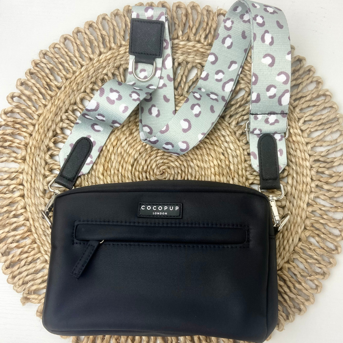 A black handbag featuring a front zipper pocket and the "Cocopup London" brand label, accessorized with an attached strap from the Sage Leopard Collection that showcases a white background with gray and purple leopard print. The bag is displayed on a circular woven mat.