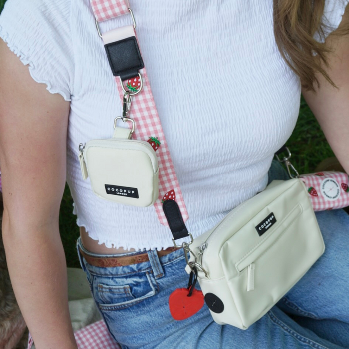 Woman in white top and jeans with two cream Cocopup London bags with strawberry-patterned straps, sitting on grass.