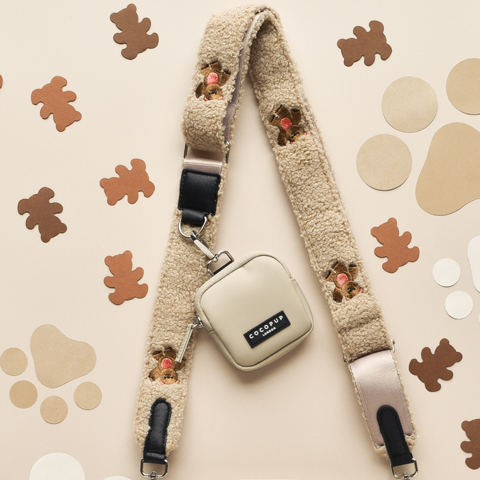 A cream-colored fuzzy camera strap from Cocopup London, known as the "Bag Strap - Theo Embroidered Teddy," featuring teddy bear face designs, is laid flat on a beige surface. Attached to the strap is a small, square, beige Dog Walking Bag with a black "ECO COUP" label. Various brown and beige paper cutouts in different shapes are placed around the strap.