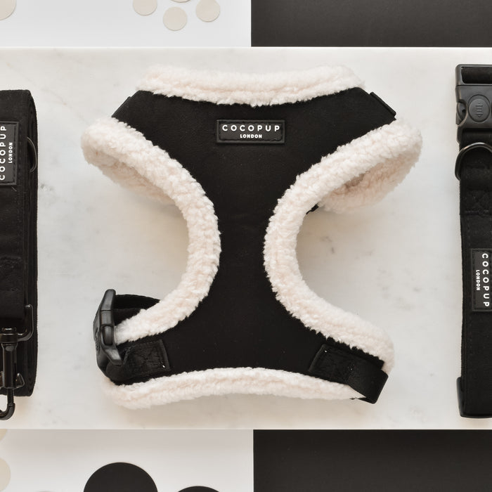 A Black Aviator Adjustable Neck Harness, Lead & Collar Bundle from Cocopup London is placed on a marble surface. The adjustable neck harness features a black and white fluffy design. To the left, the matching black leash is displayed, while to the right lies the black collar, both adorned with Cocopup London branding. The background showcases a stylish black and white polka dot theme.