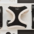 A soft, black-and-cream Black Aviator Adjustable Neck Harness from Cocopup London sits on a marble surface, accompanied by a matching black leash and collar. The items are arranged neatly and appear stylish and well-crafted, with a polka dot and black-and-white design visible in the background.