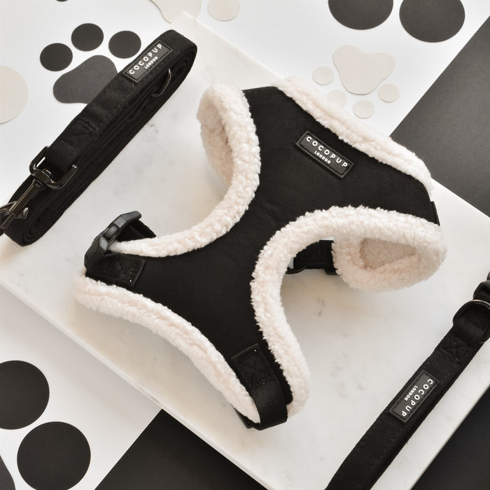 A stylish black fleece-lined Black Aviator Adjustable Neck Harness from Cocopup London is displayed on a marble surface. Two matching black leashes lie beside the harness, which features a fully adjustable chest strap. The background showcases various black and white dog paw prints.