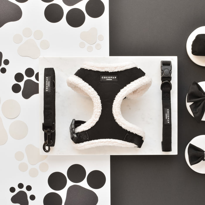 A flat lay image of Cocopup London's Black Aviator Adjustable Neck Harness, along with matching black and white dog accessories including a leash and collar, all arranged neatly on a white marble tray. The background features black and white paw print designs, with two round containers adorned with black bows placed to the right.