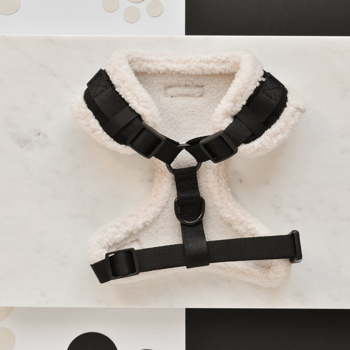 A cozy-looking, stylish dog harness from Cocopup London is placed on a marble surface. The Black Aviator Adjustable Neck Harness features a soft, cream-colored, wool-like material and is secured with black straps and buckles. With an adjustable neck harness and a fully adjustable chest strap, it includes a D-ring for leash attachment. The background is a mix of white and black surfaces.