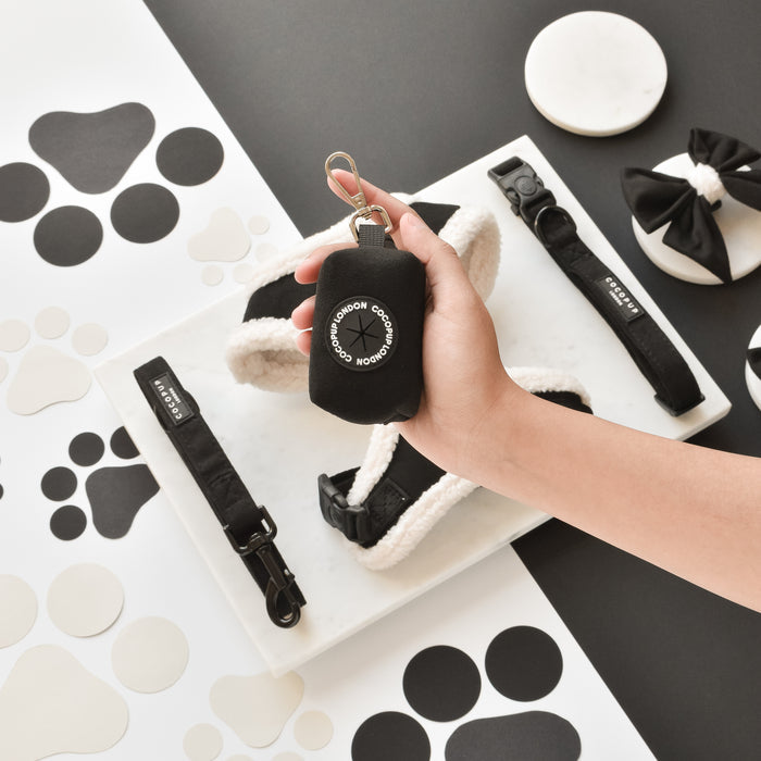 Hand holding a black Cocopup Aviator poop bag holder with matching accessories and dog poop bags on a tray.