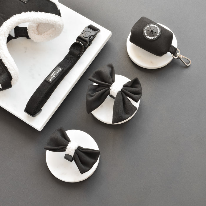 Black and white dog harness, collar, sailor bow tie, and waste bag holder set on a gray background.