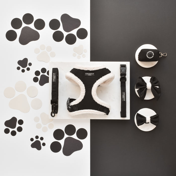 Black aviator-style sailor bow tie, matching harness, and leash by Cocopup London on paw print cutout background.