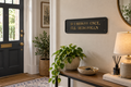 A decorative black sign with gold lettering that reads 'IF I SHENAN ONCE, I'LL SHENANIGAN' mounted on a beige wall in a styled entryway. A potted green plant, a wooden console table with a lamp, books, and a bowl sit below, with a dark front door and potted plant in the background.