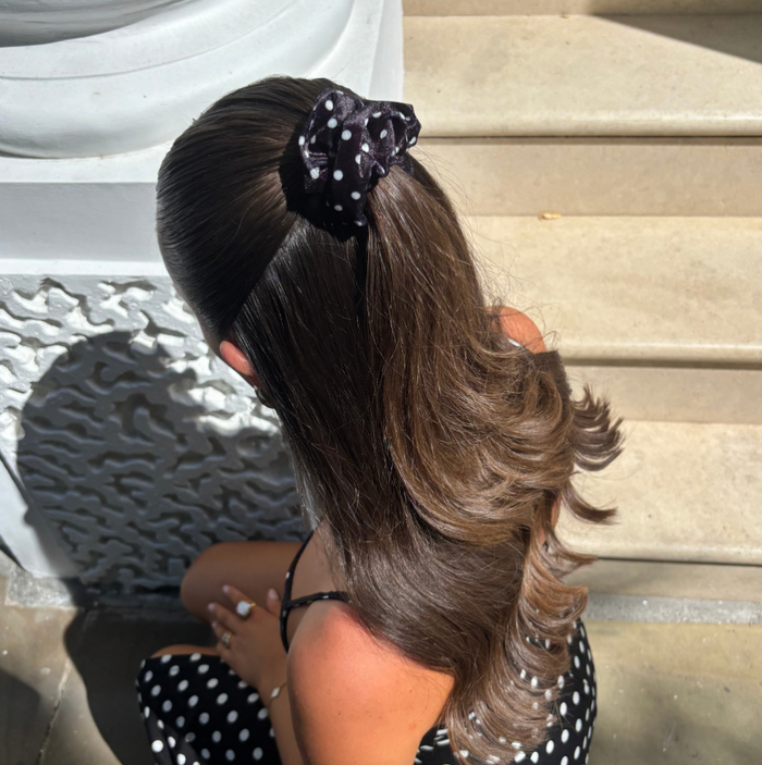 Woman with wavy brown hair wears a black polka dot scrunchie, sitting on outdoor steps.
