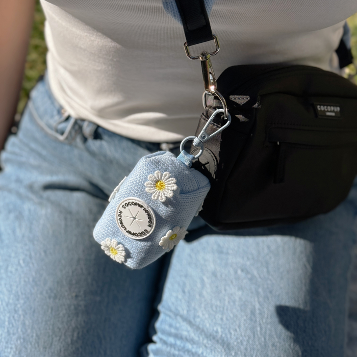 Blue Daisy Dream poop bag holder from Cocopup London attached to a black bag. Person wears jeans and a white top.
