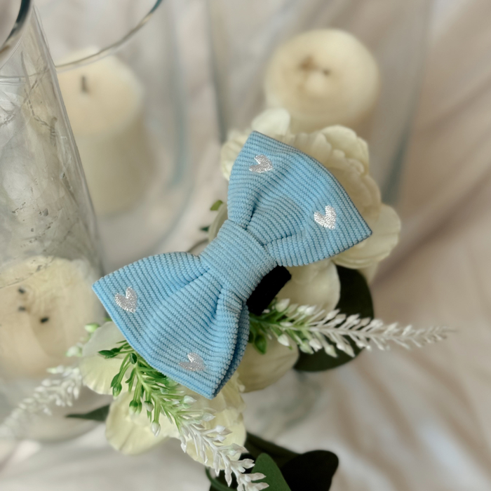 Blue cord bow tie with silver heart pattern shown on a backdrop of white flowers, candles, and glass.