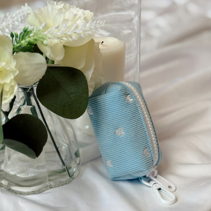 Blue cord Heartstrings poop bag holder by Cocopup London on white fabric, with flowers and a candle nearby.