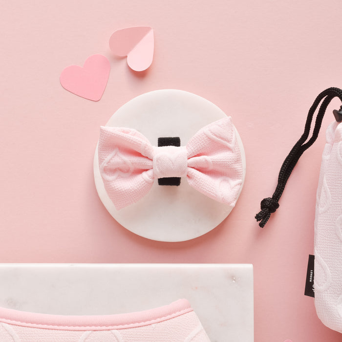 Baby pink heart-patterned bow tie by Cocopup London on a white circle, with hearts and accessories on pastel pink background.
