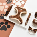 The Cocopup London Brown Aviator Adjustable Neck Harness, Lead & Collar Bundle is a stylish set featuring cream fleece lining and includes a matching leash, bow tie, and poop bag holder. The items are showcased on a white marble surface, surrounded by paper paw prints in black, brown, and beige on a brown background.