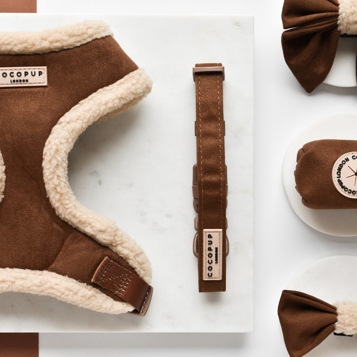 This image displays a set of brown dog accessories with labels marked "Cocopup London." The Brown Aviator Adjustable Neck Harness, Lead & Collar Bundle includes an adjustable neck harness with white fleece lining, a collar, and matching bows arranged neatly on a white marble surface.