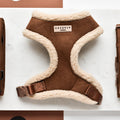 A stylish "Brown Aviator Adjustable Neck Harness" by Cocopup London, with a soft, fluffy lining and adjustable straps, is displayed on a white surface. The harness features a label that reads "COCOPUP LONDON" and has an easy-release buckle. Two additional straps or accessories are placed beside this comfortable pet harness.