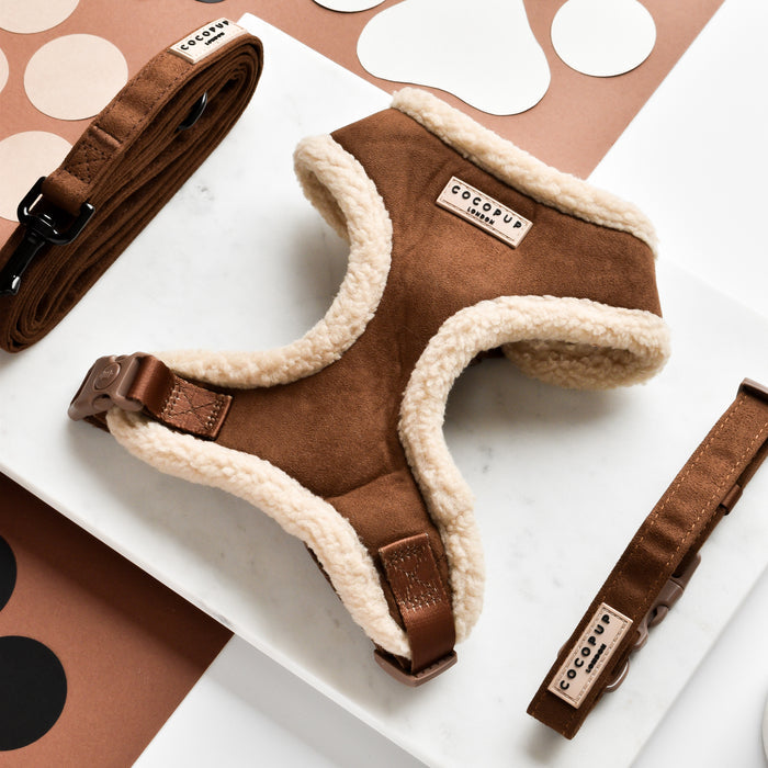 A Brown Aviator Adjustable Neck Harness from Cocopup London, featuring a stylish brown and beige design with a fuzzy lining, is displayed on a marble surface. Surrounding it are matching brown leashes and various beige and black shapes set against a brown background. The comfortable pet harness and leashes have labels with the brand name “Cocopup London” on them.