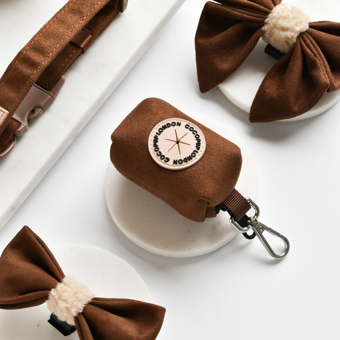 Brown Aviator poop bag holder by Cocopup London with faux suede bows and collar, shown on a white marble surface.