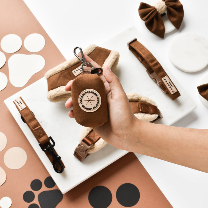 Hand holding a brown Cocopup London Aviator poop bag holder, with matching dog accessories on marble and brown surfaces.