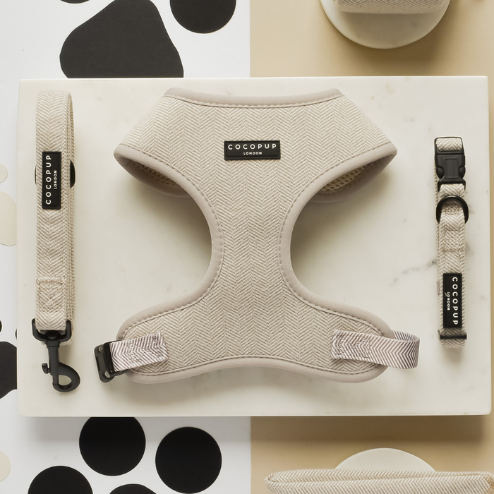 A stylish dog harness set displayed on a white background with geometric patterns. The Brown Tweed Adjustable Neck Harness, Lead & Collar Bundle by Cocopup London includes a beige herringbone fabric harness, leash, and collar, all featuring black clasps and "Cocopup London" branding labels. Perfect for chic dog walks.